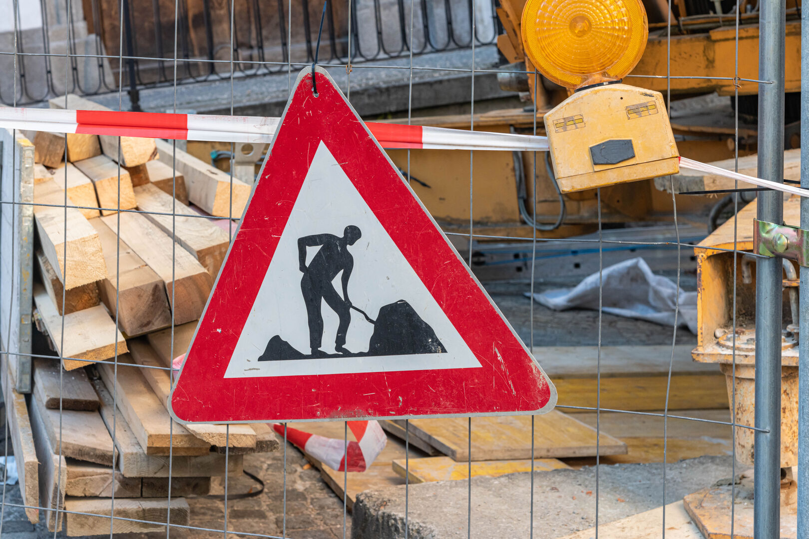 Baustellenschild mit Mann, der schaufelt, vor Holzstapel und Absperrgittern